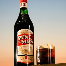 Load image into Gallery viewer, Bottle of Punt e Mes aperitivo with a glass filled with the drink against a sunset sky.