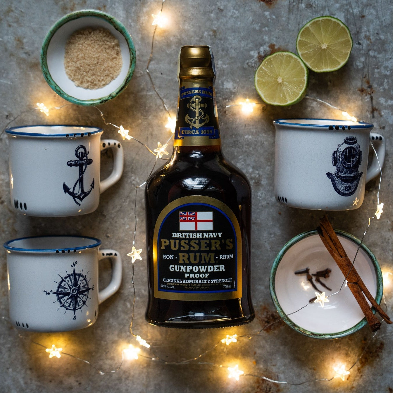 Load image into Gallery viewer, Bottle of Pusser’s Gunpowder Proof Rum surrounded by mugs, limes, and spices on a rustic surface with string lights.