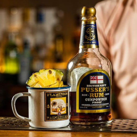 Load image into Gallery viewer, Bottle of Pusser’s Gunpowder Proof Rum with a pineapple and mug on a bar counter, blurred bar background