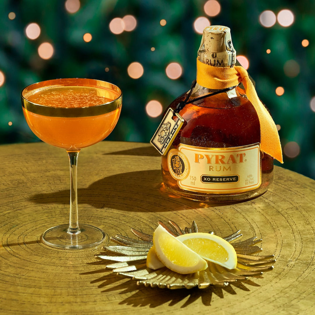 Load image into Gallery viewer, Pyrat XO Reserve Rum bottle with a cocktail and lemon slices on a wooden surface with a festive background.