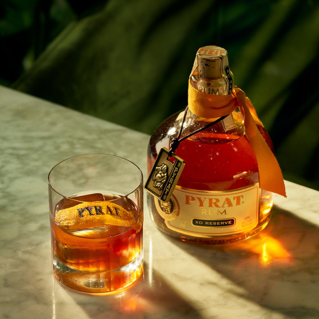 Load image into Gallery viewer, Pyrat XO Reserve rum bottle and glass on a reflective surface with greenery in the background