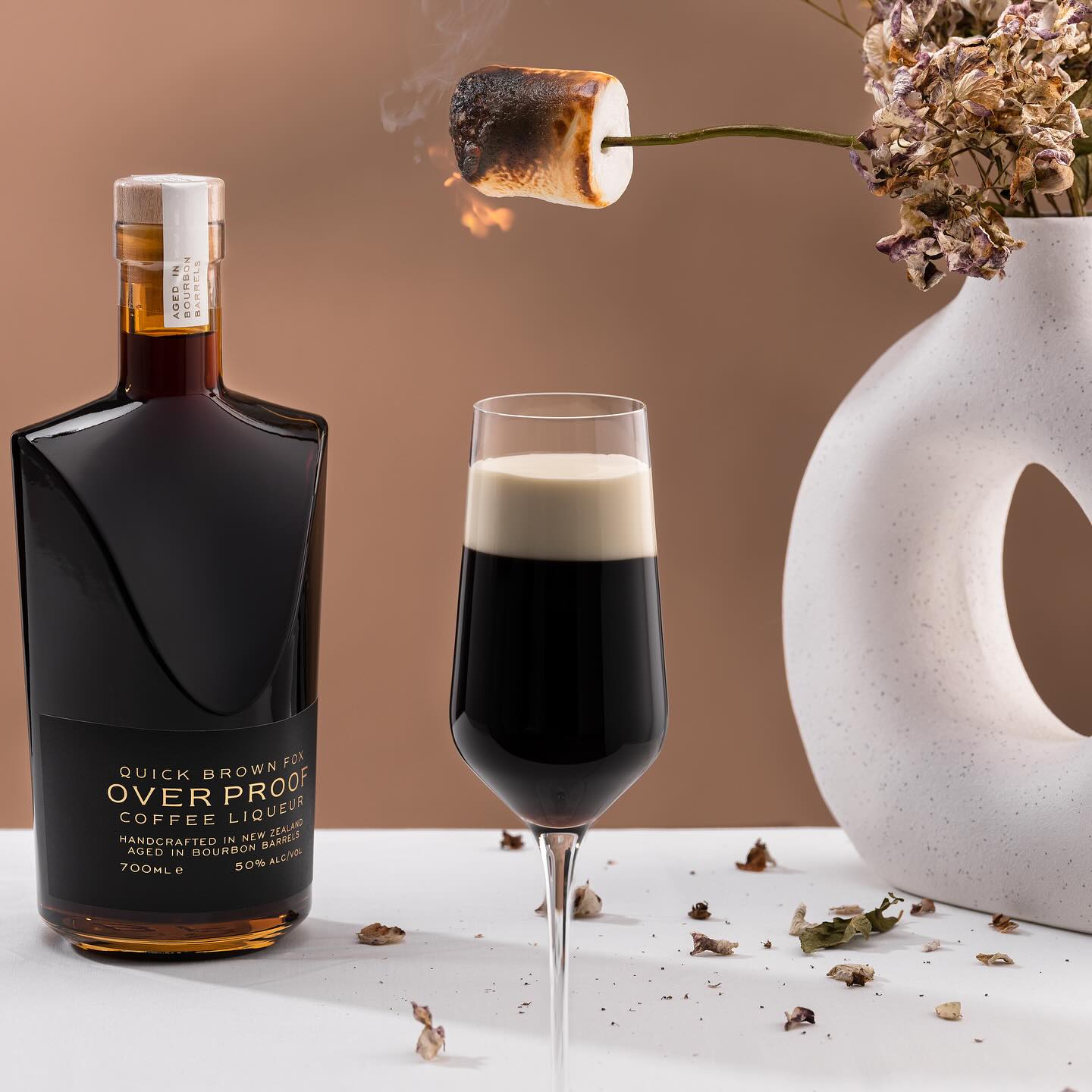 Load image into Gallery viewer, A bottle of Quick Brown Fox Coffee Liqueur with a coffee drink in a champagne glass on a table and some dried flowers in a vase