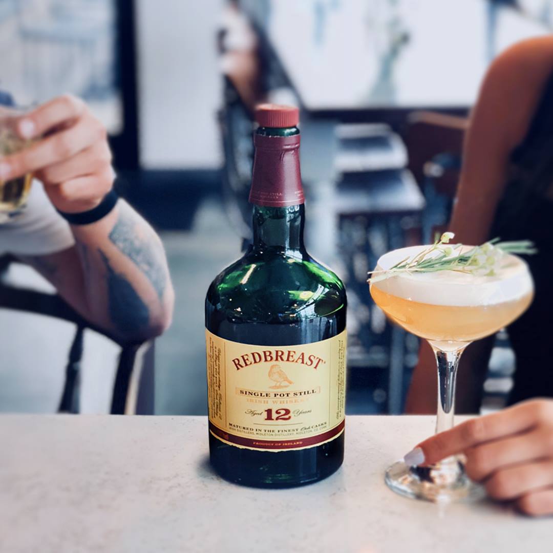 Load image into Gallery viewer, Redbreast 12 year old whiskey bottle on a table with a cocktail in the background