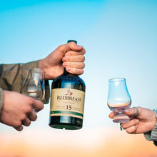 Load image into Gallery viewer, Two people toasting with a bottle of Redbreast 15 year old whiskey and two glasses against a blue sky.