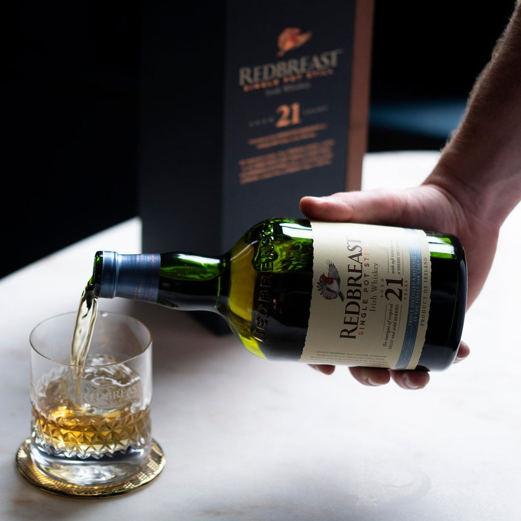 Person pouring Redbreast 21 YEAR OLD whiskey into a glass with a bottle and box in the background.