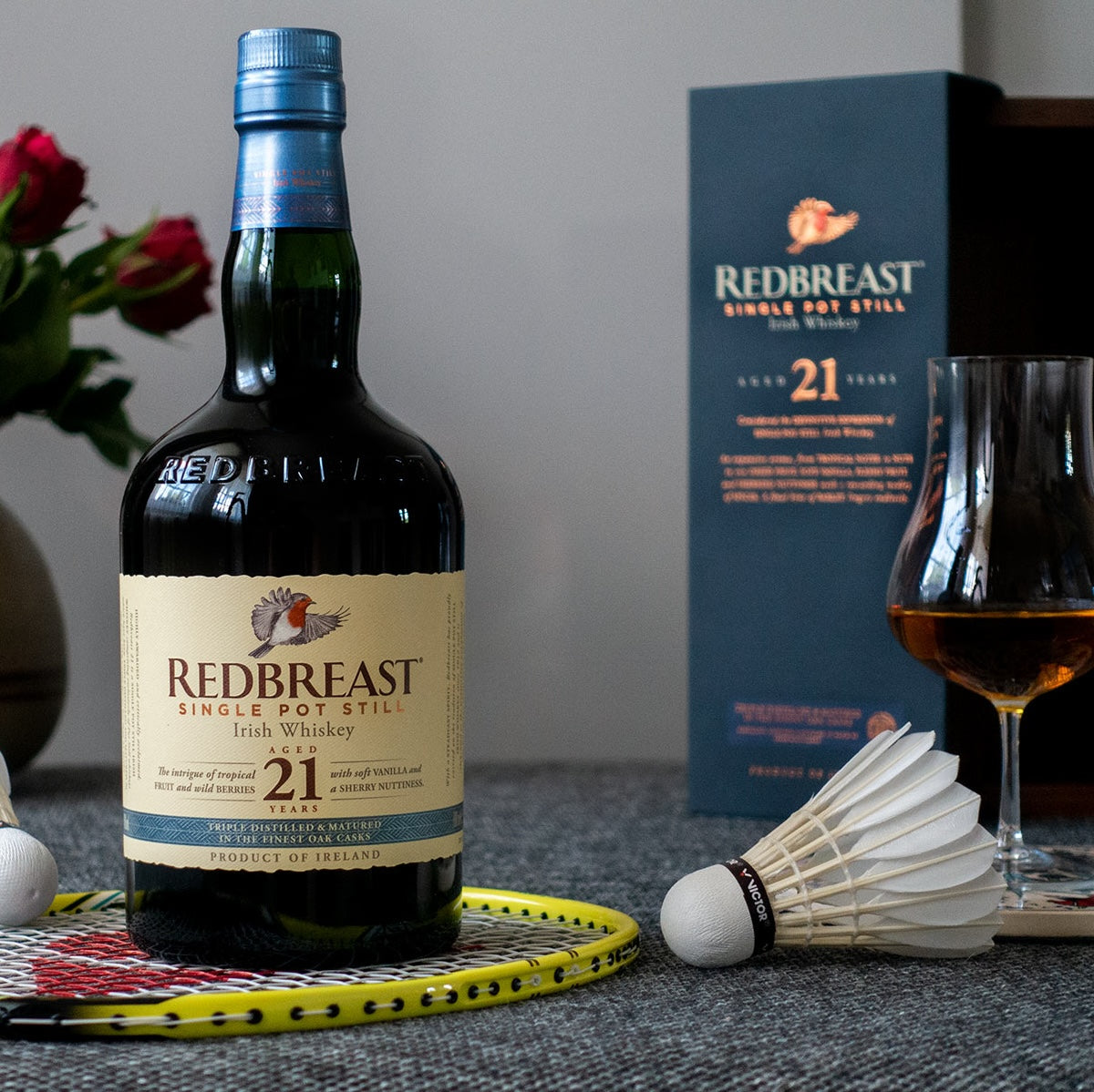 Load image into Gallery viewer, A bottle of Redbreast 21YO Irish Whiskey standing on a badminton racket with the ball beside it and a glass of neat whiskey in it