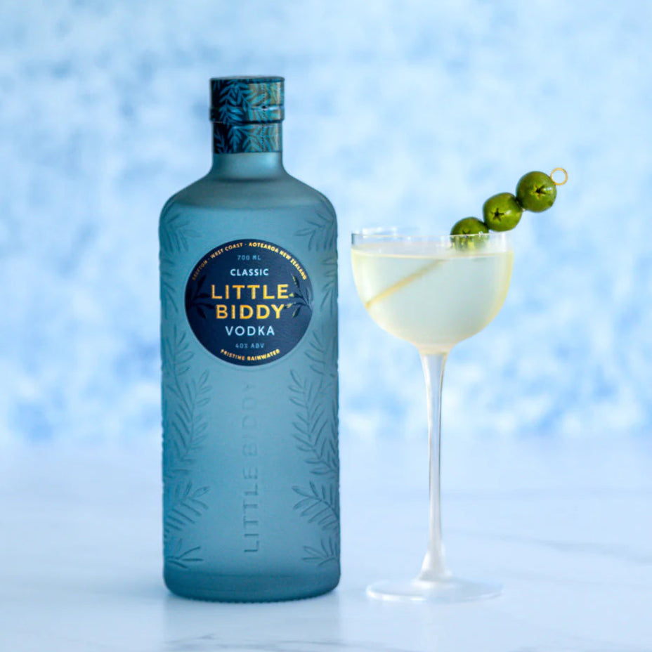 Bottle of Little Biddy Vodka with a cocktail garnished with green olives on a light blue background