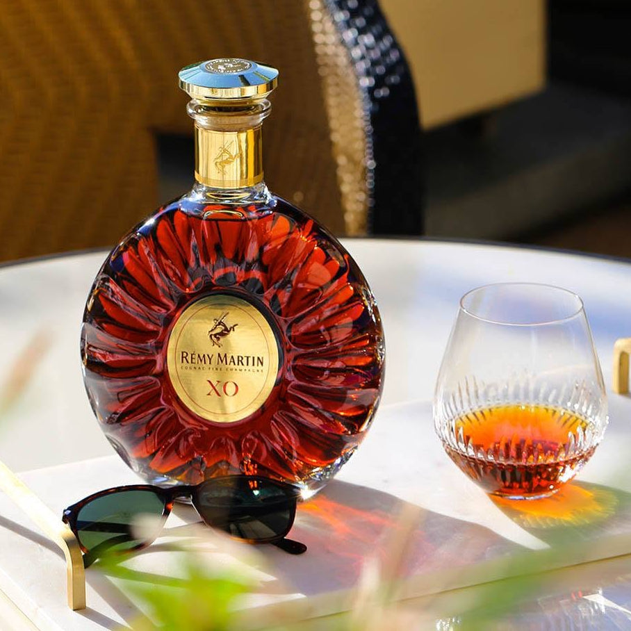 Load image into Gallery viewer, A bottle of Remy Martin XO cognac with a gold label and blue cap, displayed on a white tray next to a glass of the amber liquid, with sunglasses in the foreground.