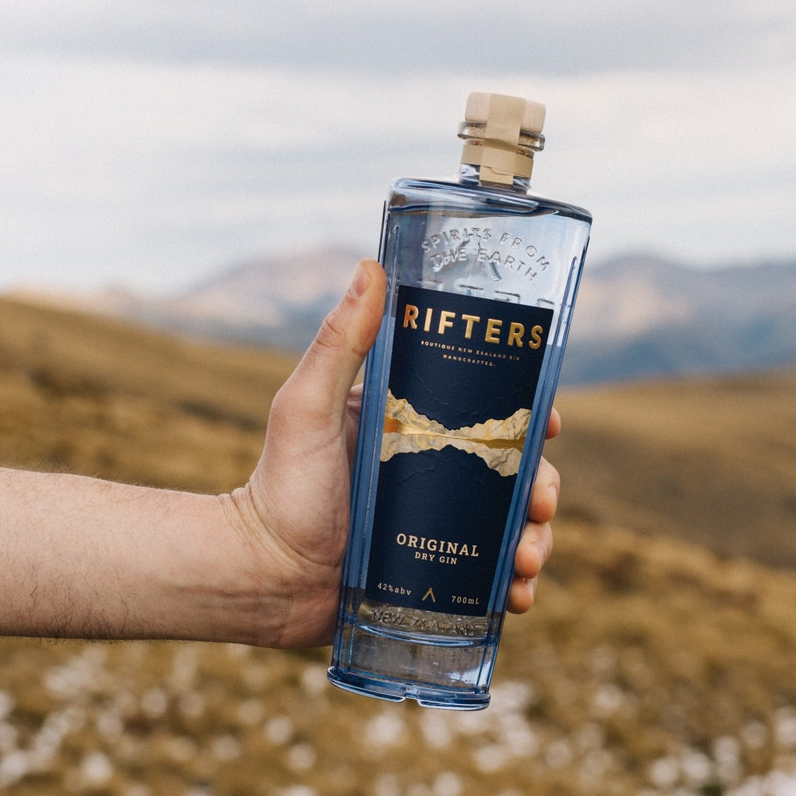 Load image into Gallery viewer, Hand holding a bottle of Rifters Original Dry Gin with a scenic background
