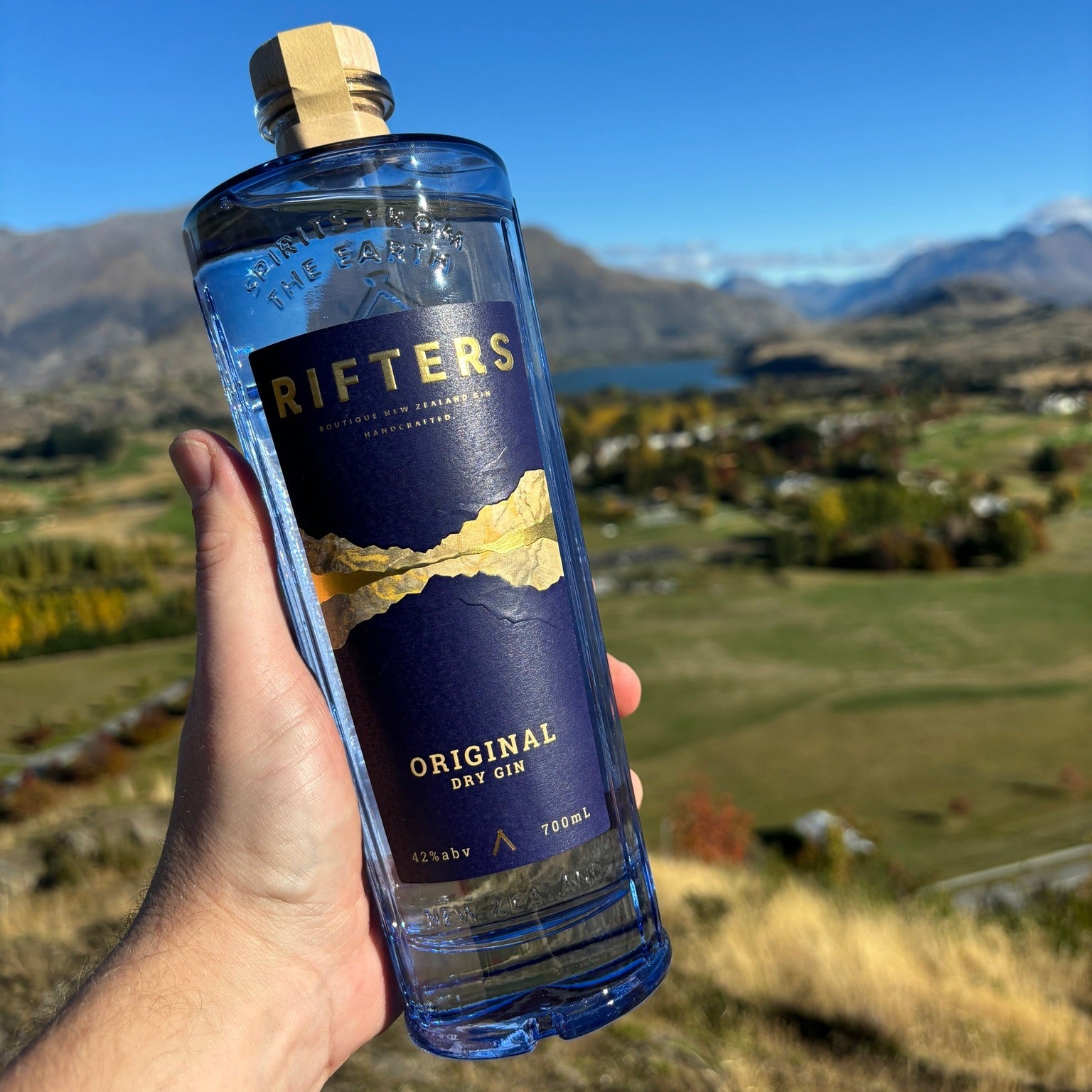 Load image into Gallery viewer, Bottle of Rifters Original Dry Gin held in front of a scenic landscape with mountains and a lake.