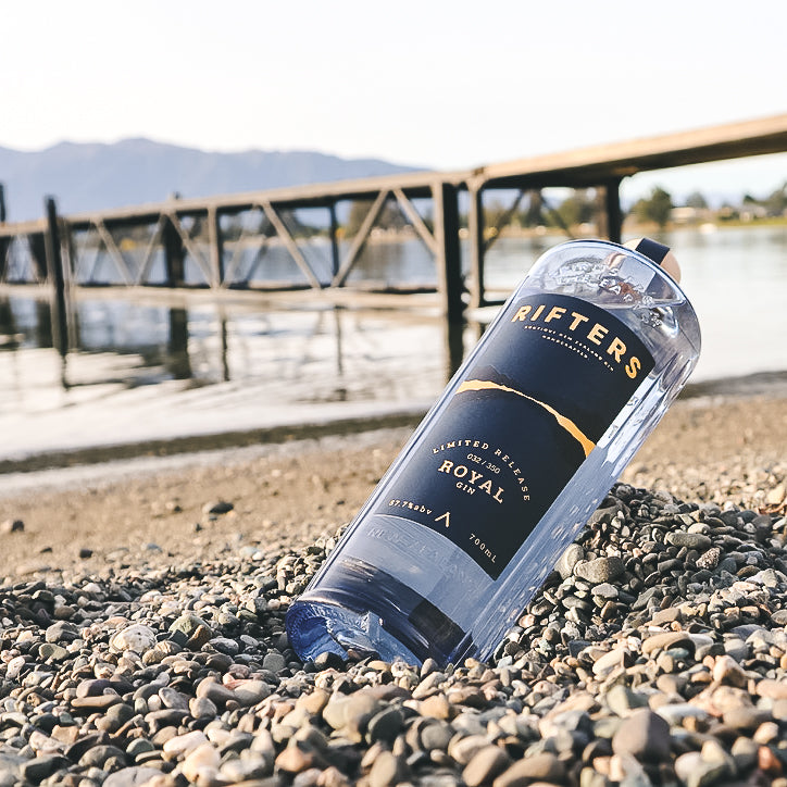 Load image into Gallery viewer, Bottle of Rifters Royal Navy Strength Gin lying on pebbles by a waterfront with a bridge in the background