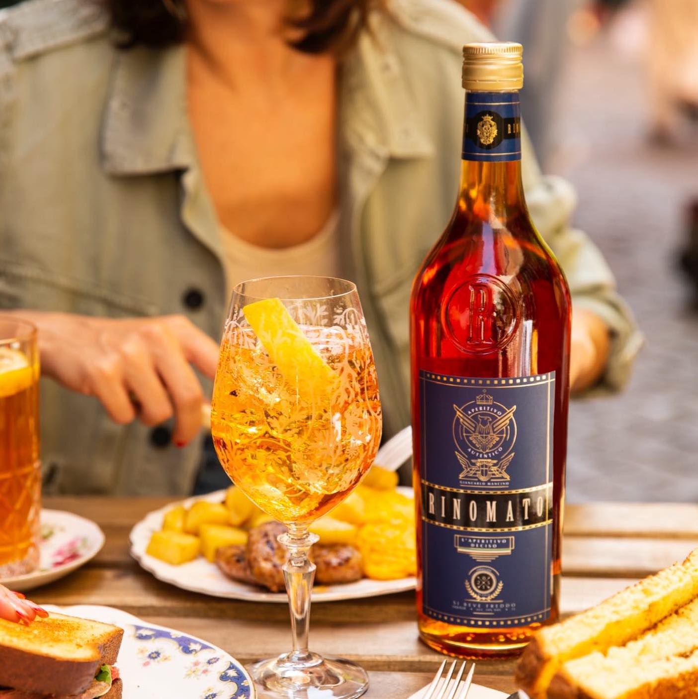Load image into Gallery viewer, A bottle of Rinomato L'Aperitivo and cocktail with a lemon wedge on a table with food and a woman in the background eating 