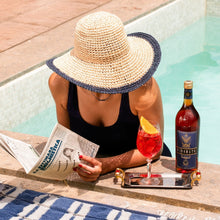 Load image into Gallery viewer, Person by a pool reading a newspaper with a bottle of Rinomato nd a cocktail.