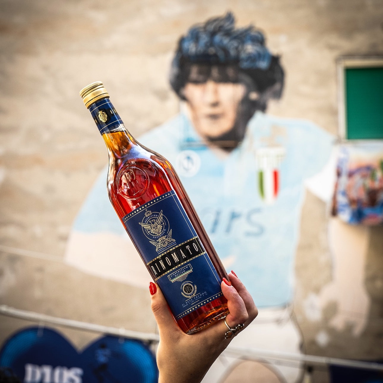 Load image into Gallery viewer, A hand holding a bottle of Rinomato with a blurry background of a painting of an Italian football player