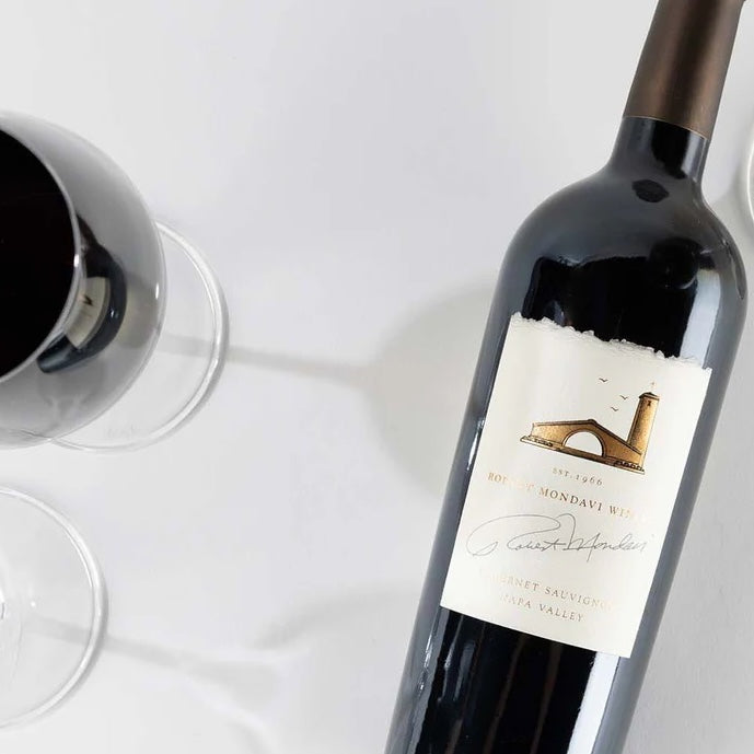 Load image into Gallery viewer, A close up image of a bottle of Mondavi Napa Cabernet Sauvignon red wine next to a half full wine glass taken from above.