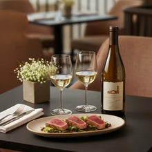 Load image into Gallery viewer, Wine bottle, two glasses of white wine, and a plate of food on a table in a restaurant setting.