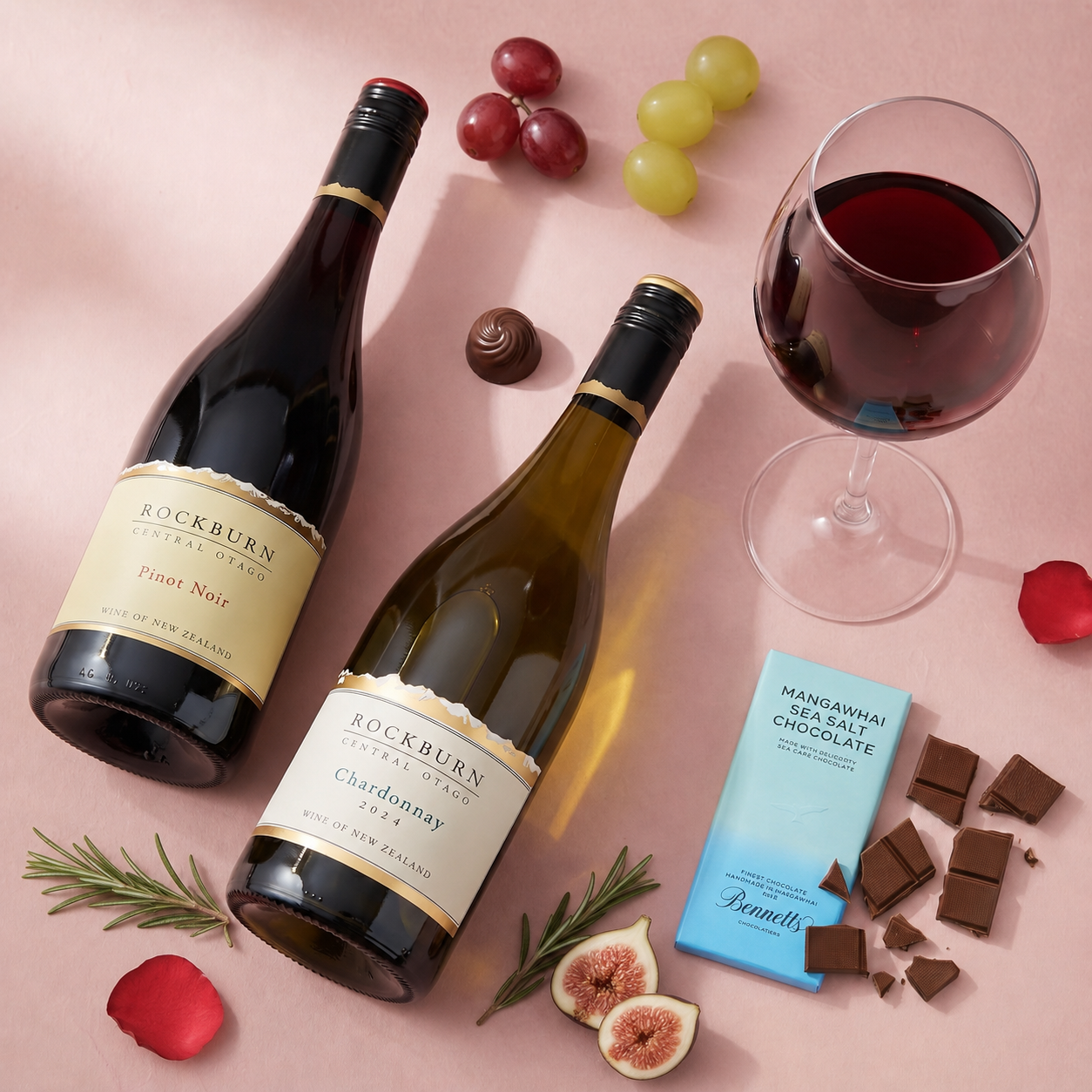 Load image into Gallery viewer, Two bottles of Rockburn Winery wine, a glass of red wine, grapes, chocolate, figs, rosemary, and a blue Bennett chocolate bar, arranged on a pink background.
