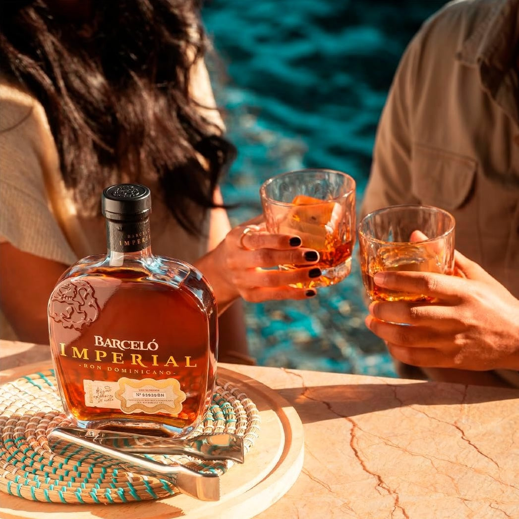 Load image into Gallery viewer, Barceló Imperial rum bottle with two glasses of orange liquid by a poolside.