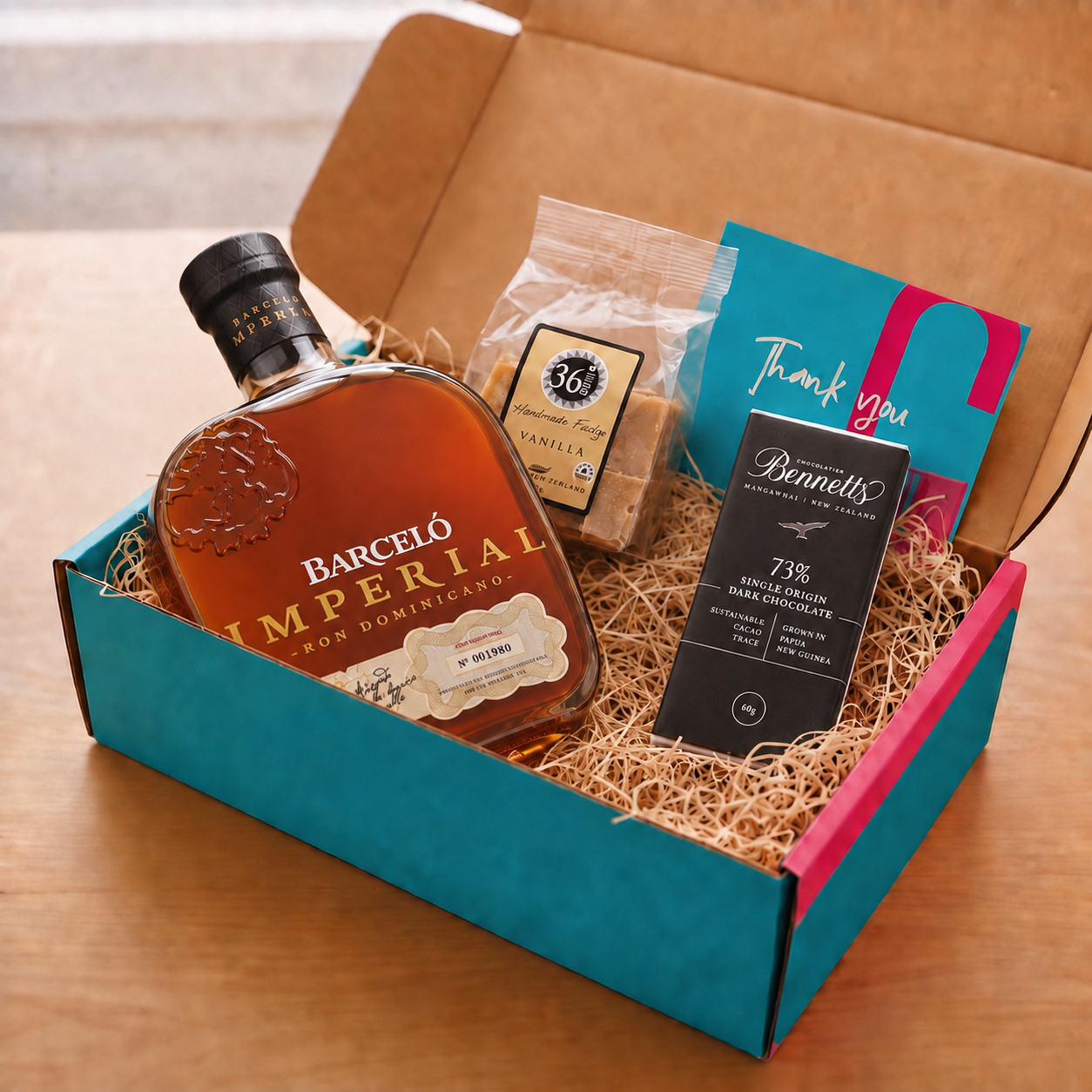 Load image into Gallery viewer, A curated gift box set on a wooden table, featuring a bottle of Ron Barceló Imperial rum, a packet of 36 Degrees Vanilla fudge, a bar of Bennetts 73% dark chocolate, and a teal and pink 'Thank you' card inside a turquoise and pink COVE 27 gift box with wood-wool.