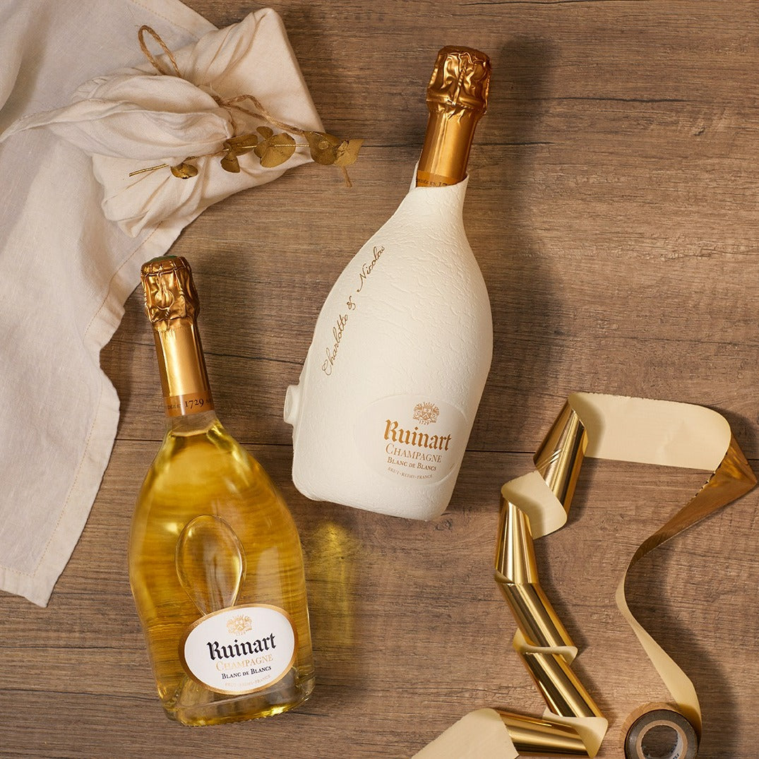 Load image into Gallery viewer, Two Ruinart champagne bottles on a wooden surface with a decorative cloth and bottle opener.