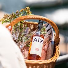 Load image into Gallery viewer, Bottle of AIX rose in a wicker basket with flowers and glasses, blurred background