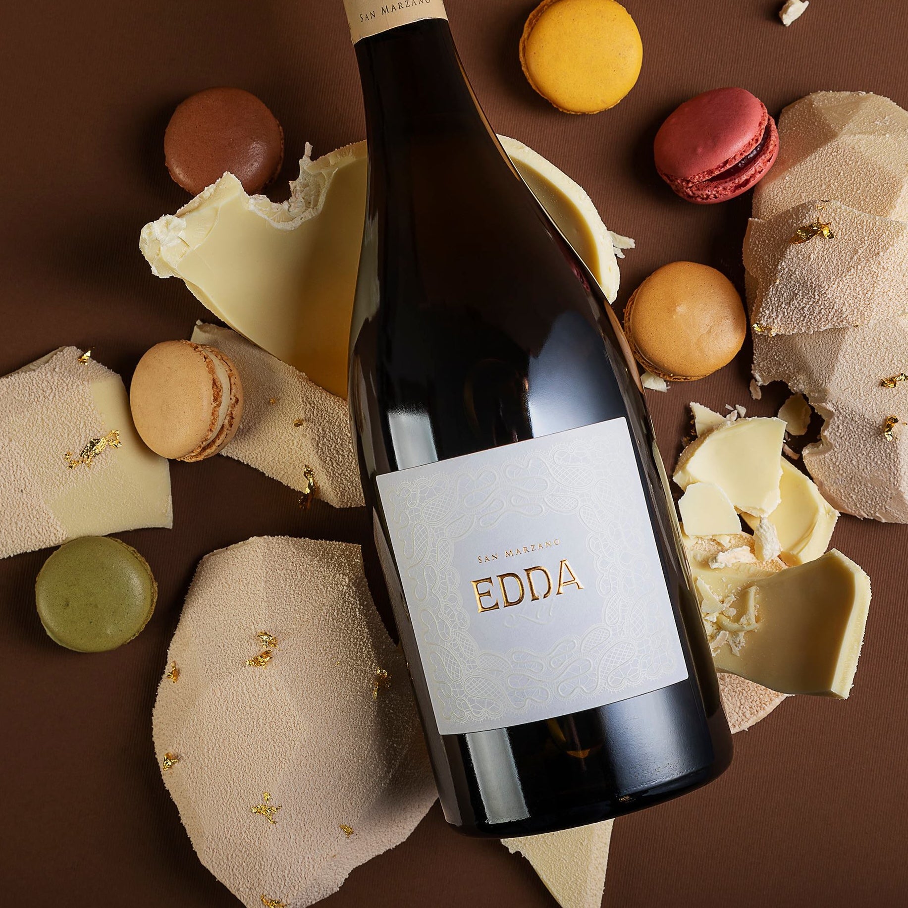 Load image into Gallery viewer, a bottle of white wine laying on coloured macaroons on a brown cloth