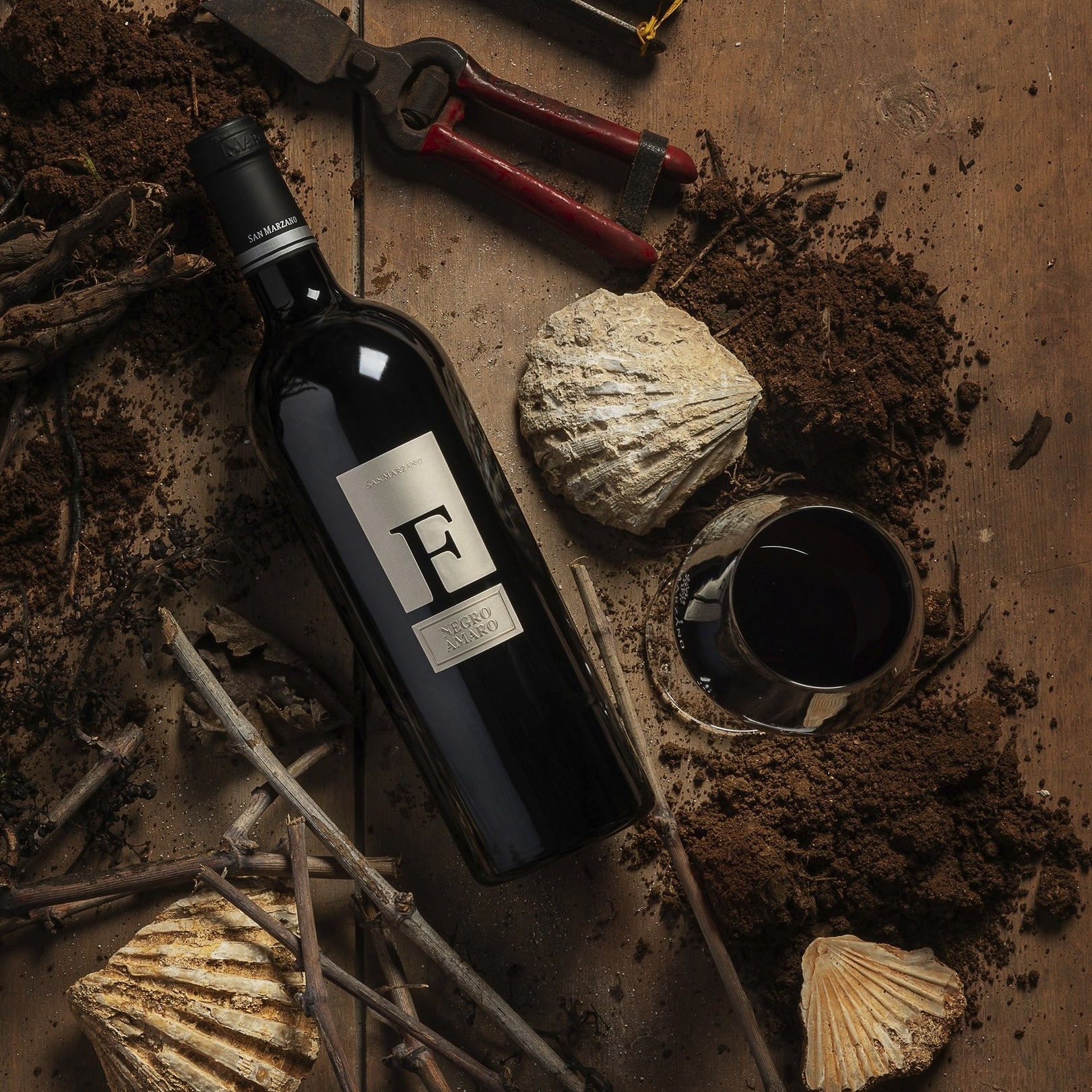Load image into Gallery viewer, A bottle of San Marzano F Puglia Negroamaro red wine on sand with shells and glass of red wine beside