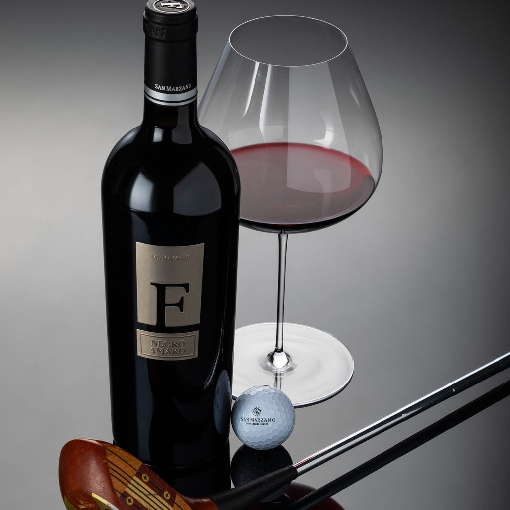 San Marzano F Puglia Negroamaro wine bottle, glass with red wine, golf club, and golf ball on a reflective surface