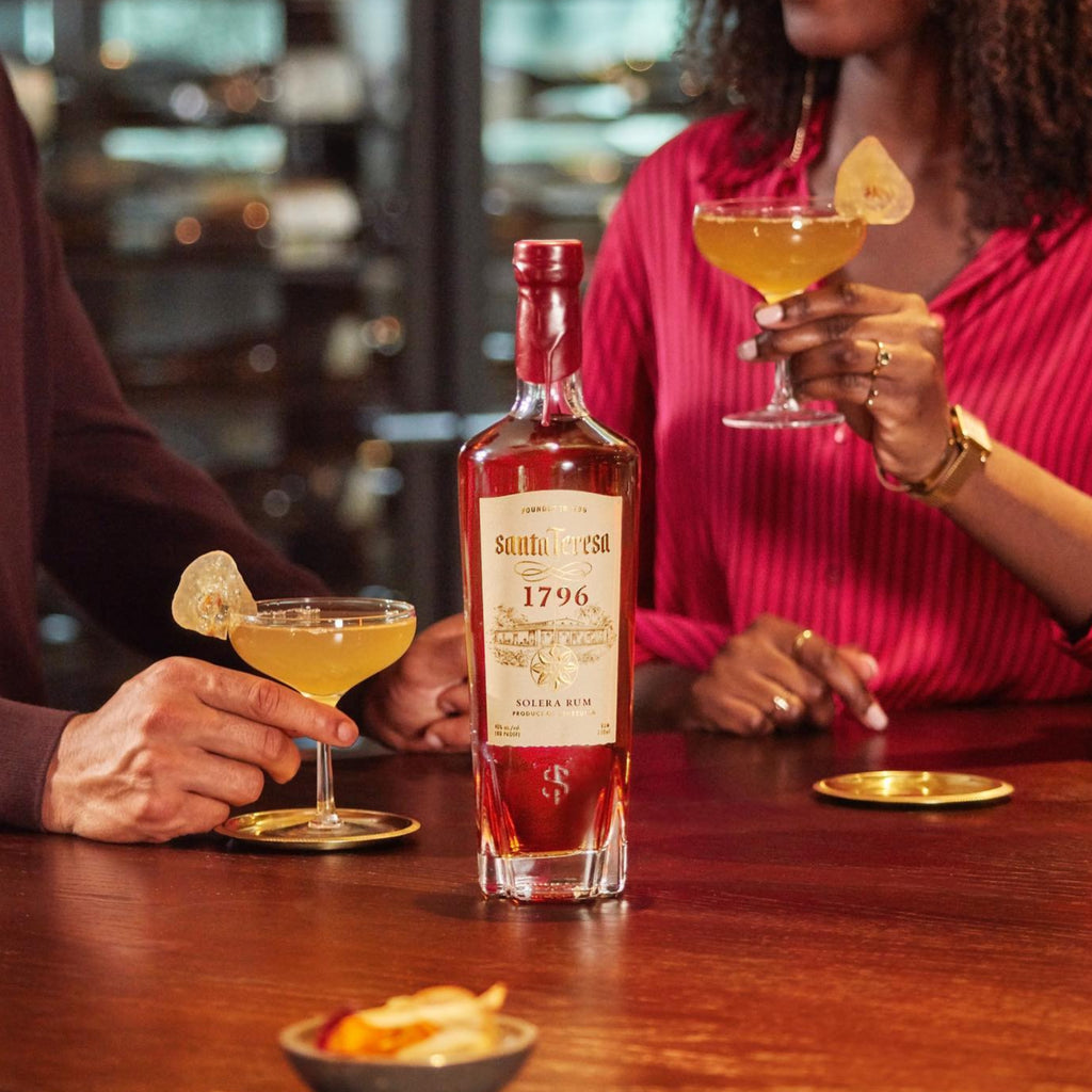 Two people at a bar holding cocktails with a bottle of Santa-Teresa-1796-Rum in the foreground.