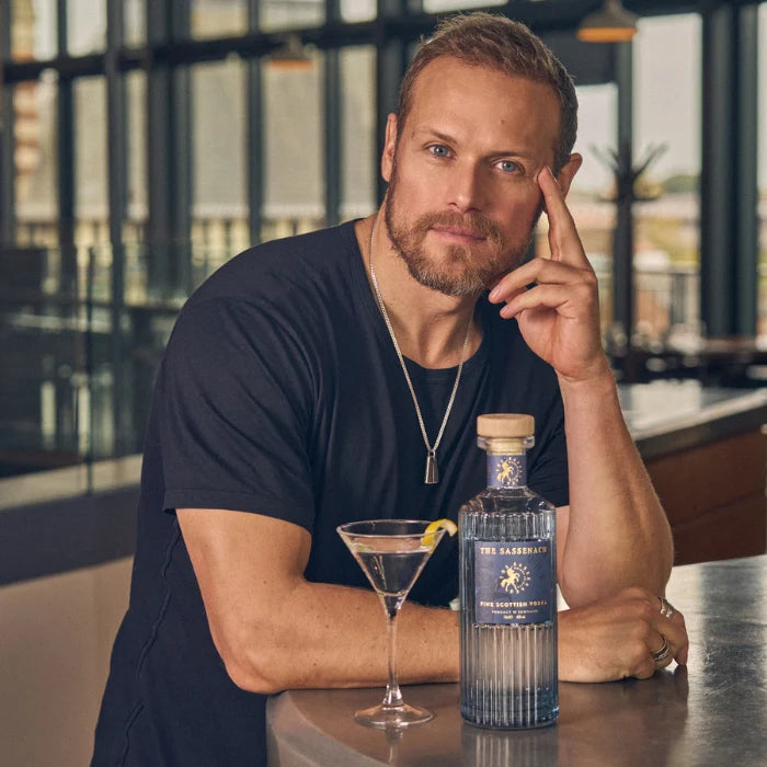 Load image into Gallery viewer, Actor Sam Heughan sitting at a bar with a bottle of The Sassenach Fine Scottish Vodka and a cocktail glass.