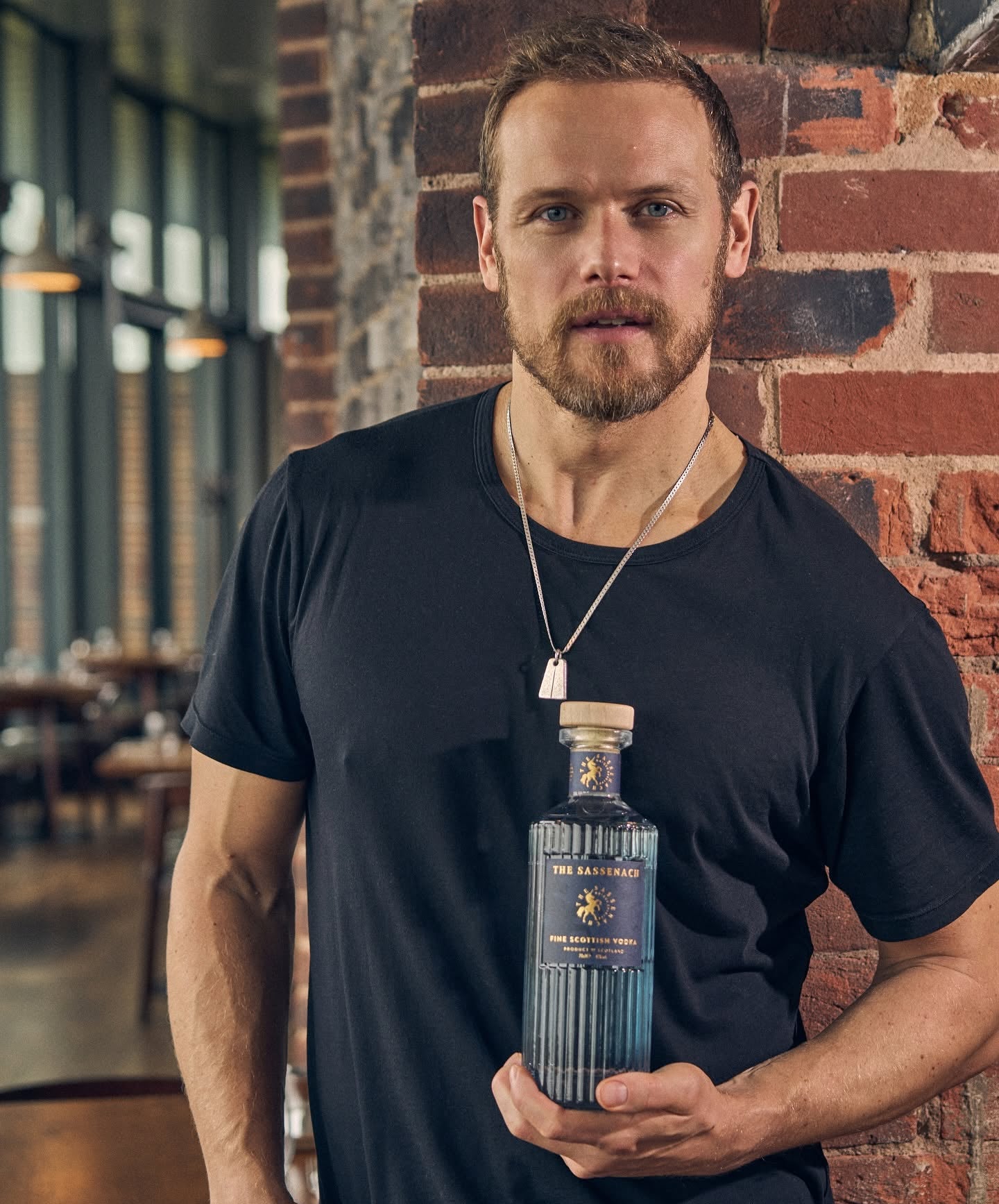 Load image into Gallery viewer, Actor Sam Heughan wearing a black t-shirt and silver necklace holds a bottle of The Sassenach Fine Scottish Vodka against a brick wall background.