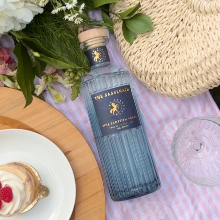 Load image into Gallery viewer, A bottle of The Sassenach Fine Scottish Vodka is styled on a pink and white striped picnic blanket with a straw bag, flowers, a dessert, and glassware.