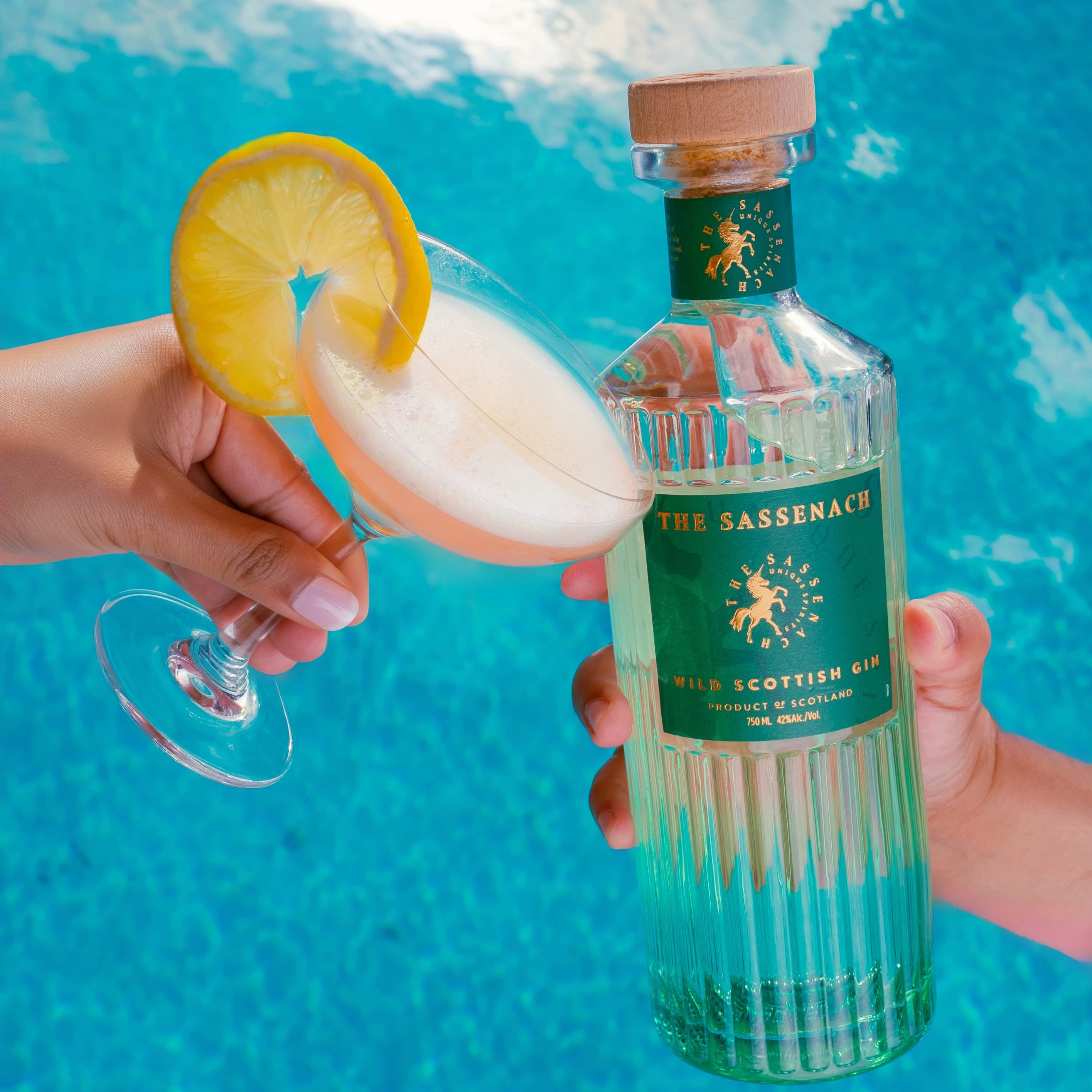 Load image into Gallery viewer, A bottle of The Sassenach Highland Scottish Gin held by hands against a bright blue swimming pool background, with a cocktail glass and lemon garnish in the foreground.