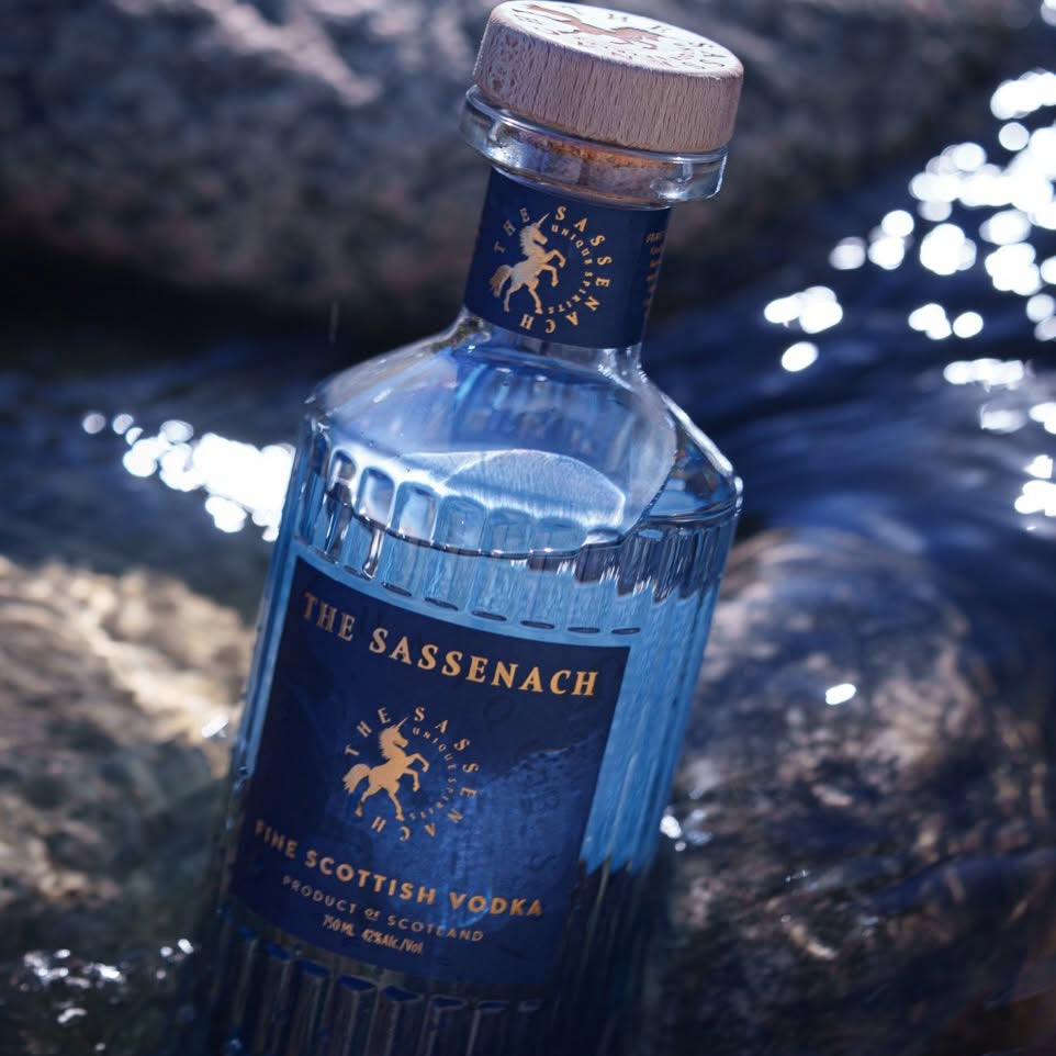 Load image into Gallery viewer, A bottle of The Sassenach Fine Scottish Vodka partially submerged in flowing water, with dark rocks and sunlight reflecting around it.