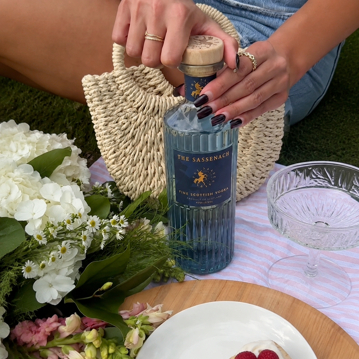 Load image into Gallery viewer, A woman outdoors at a picnic setting holds a bottle of The Sassenach Fine Scottish Vodka with a wood and cork stopper. The scene includes a woven bag, flowers, a glass, and a plate on a pink striped cloth.
