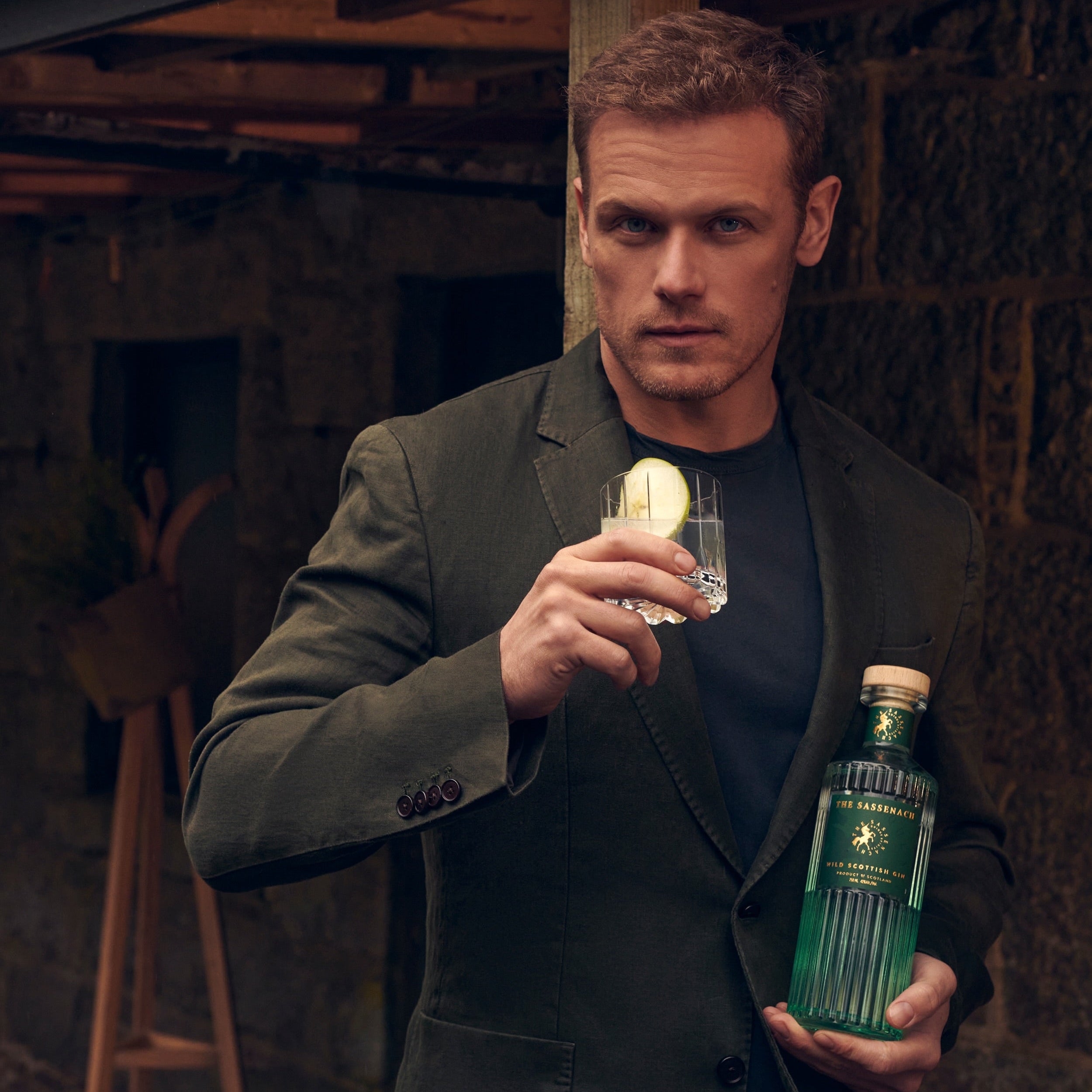 Load image into Gallery viewer, The actor Sam Heughan from the TV Series Outlander in a dark olive green blazer holds a clear cocktail glass with a lemon slice and a bottle of his Sassenach Scottish Gin, posed in a rustic stone-walled setting.