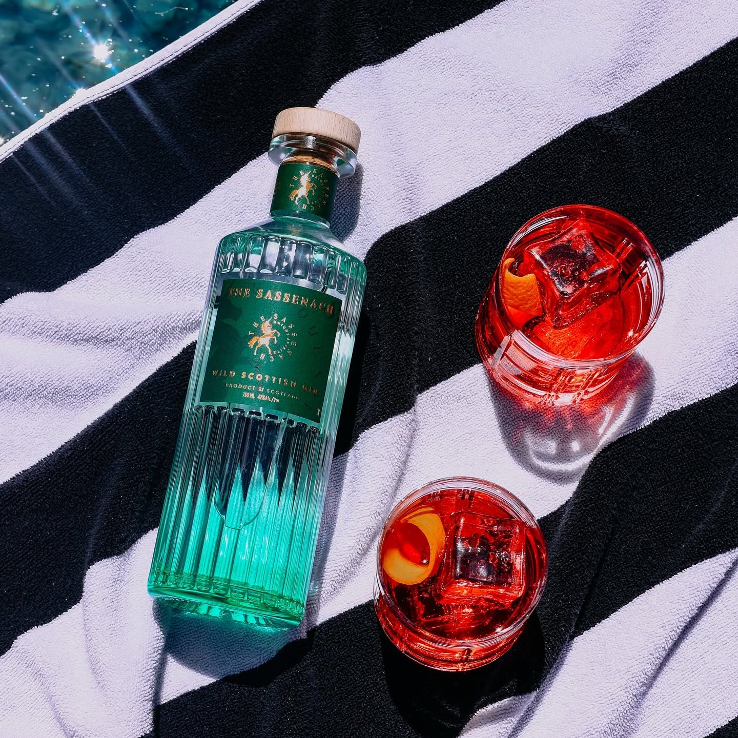 Load image into Gallery viewer, A bottle of Sassenach Scottish Gin Tonic with two filled cocktail glasses on a black and white striped towel near a pool.