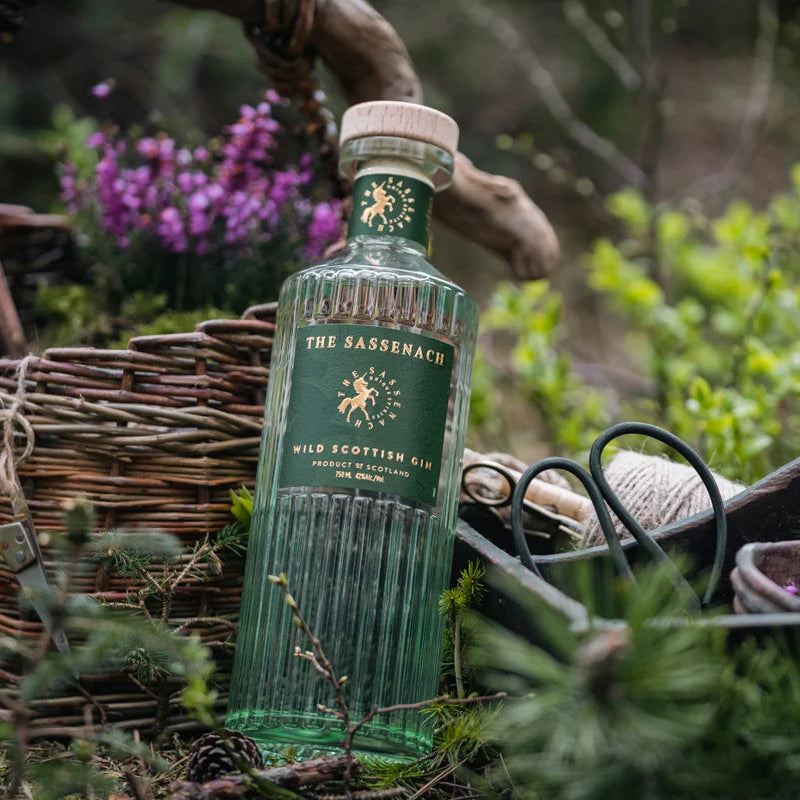 Load image into Gallery viewer, A bottle of The Sassenach Wild Scottish stands in a rustic outdoor setting with a wicker basket, gardening tools, and green foliage in the background.