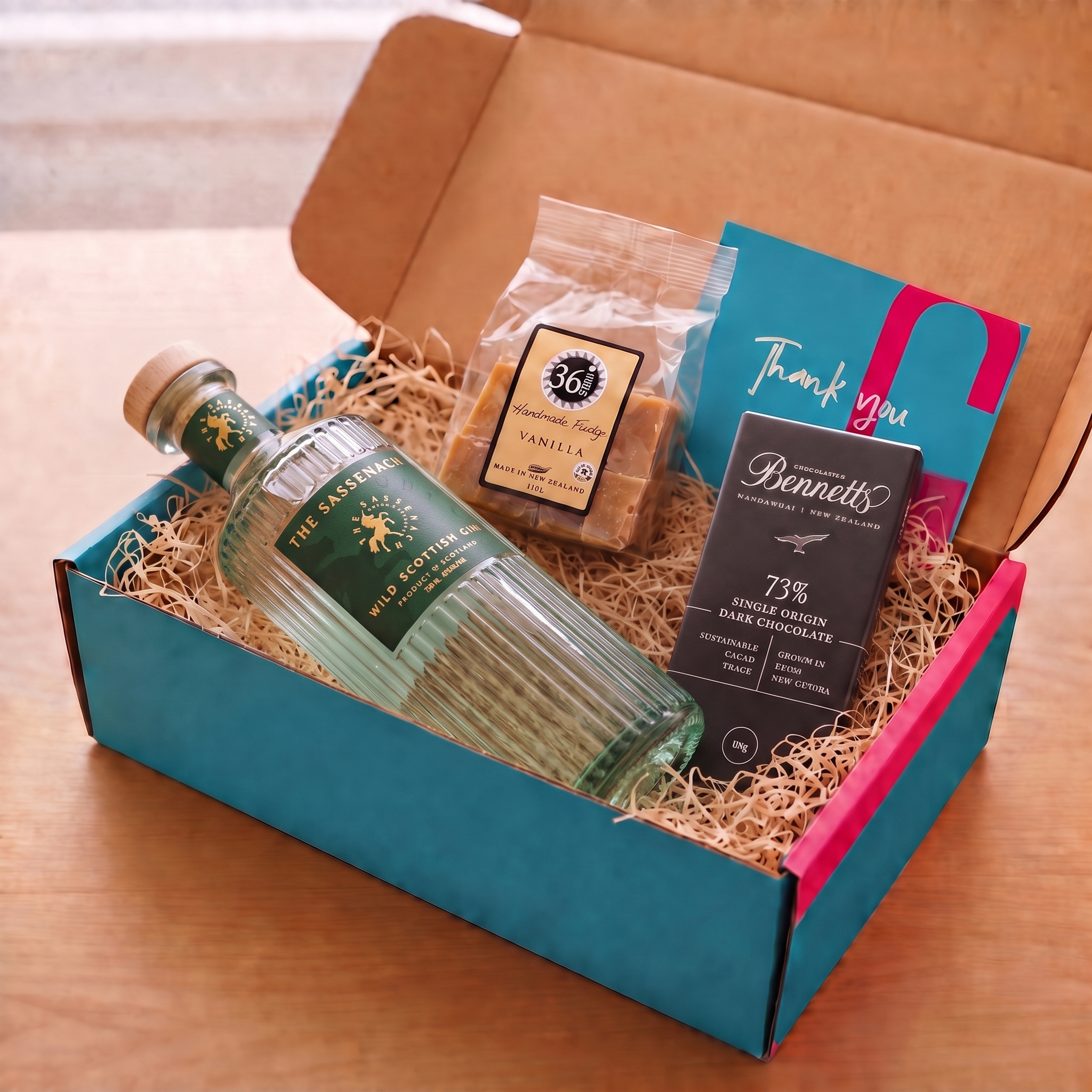 Load image into Gallery viewer, A gift box set on a wooden table containing a bottle of The Sassenach Wild Scottish, a bag of 36 Degrees Vanilla fudge, a Bennett Dark Chocolate bar, and a teal and pink Thank You card, arranged in wood-wool inside a COVE 27 branded turquoise and pink box.