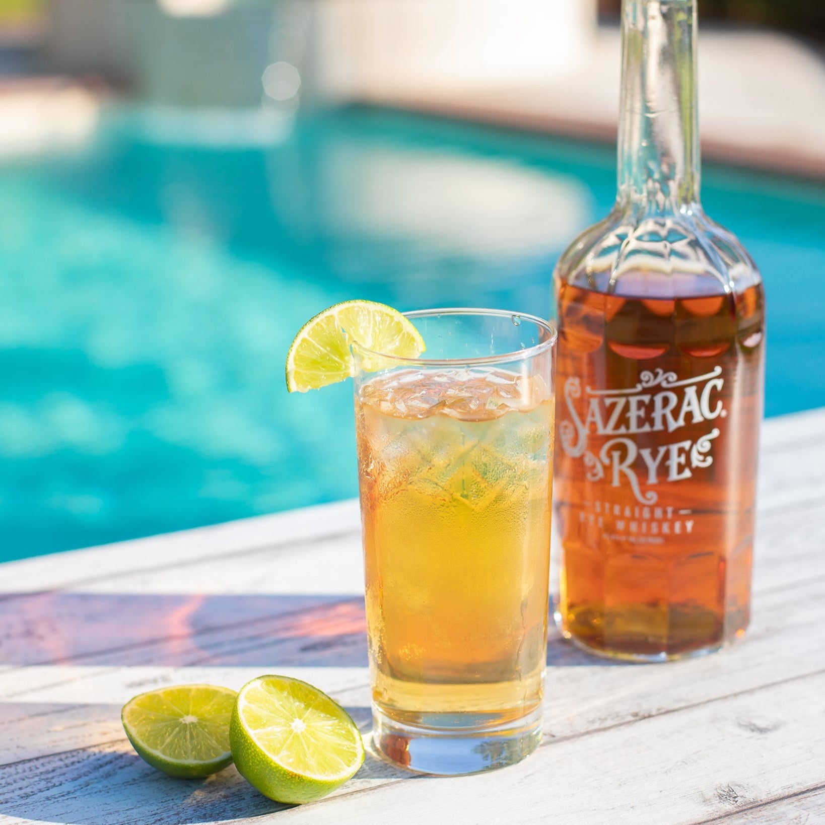 Load image into Gallery viewer, Sazerac Rye bottle with a glass of iced tea and lime wedges by a pool.