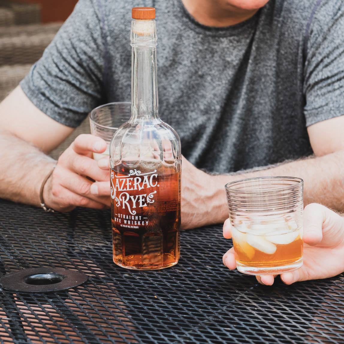 Load image into Gallery viewer, Person holding a bottle of Bazerac Rye whiskey and a glass with a drink on a metal table.