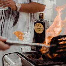Load image into Gallery viewer, Person holding a bottle of Scapegrace Gold 
gin over a fire pit with glasses nearby.