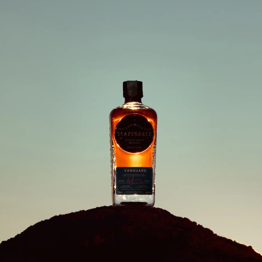 Load image into Gallery viewer, Bottle of Scapegrace Vanguard Single Malt Whisky on a small hill with a gradient sky