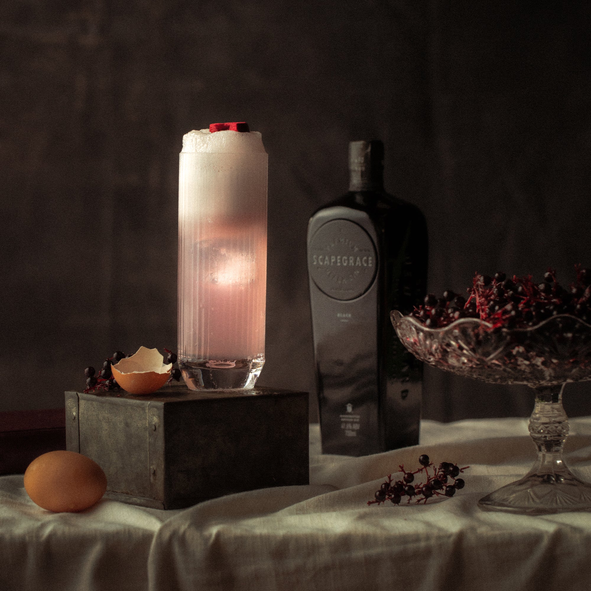 Load image into Gallery viewer, Scapegrace Black Gin bottle and a pink cocktail in a highball glass on a table with decorative items and a lit candle.