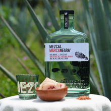 Load image into Gallery viewer, MEZCAL MARCANEGRA bottle with a small bowl of fruit and a glass on a white surface with green plants in the background