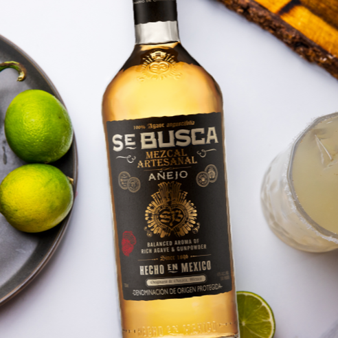 Load image into Gallery viewer, Bottle of Se Busca Mezcal with limes and a jalapeño on a plate, with a cocktail in the background.