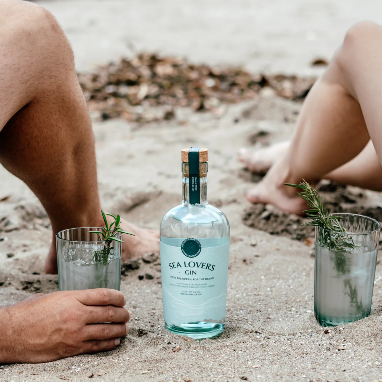 Load image into Gallery viewer, Two people sitting on a beach with a bottle of gin and a couple of glasses of gin and tonics.