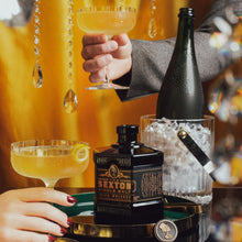 Load image into Gallery viewer, Two people holding cocktails with a bottle of Sexton Irish whiskey on a tray against a yellow curtain background.