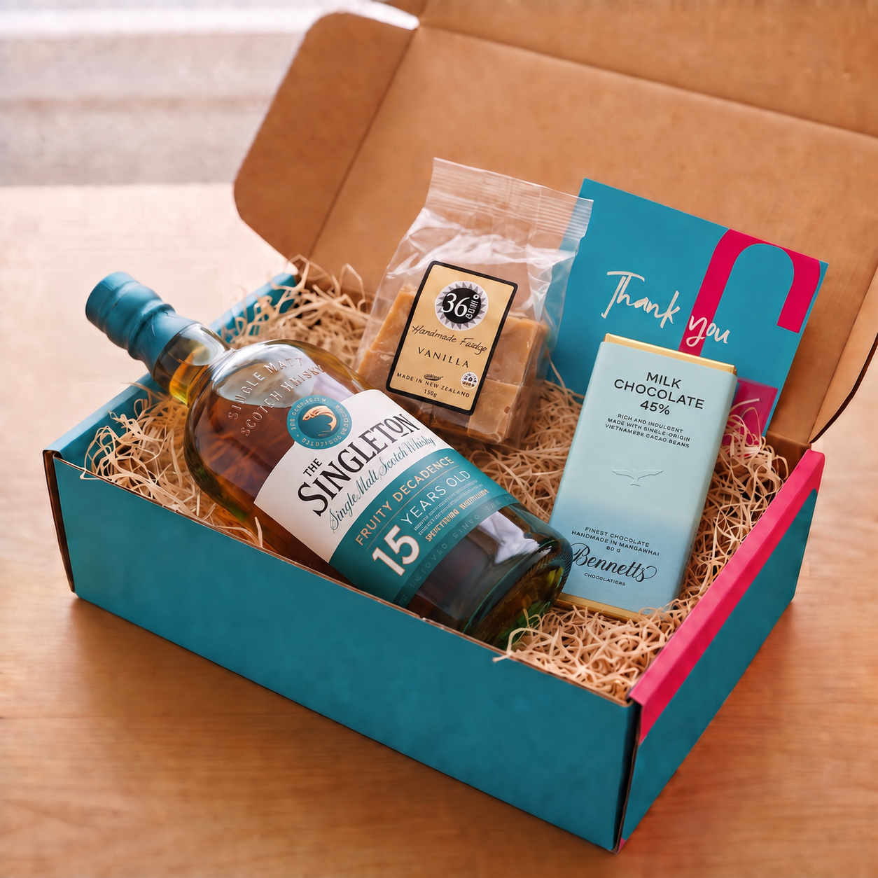 Load image into Gallery viewer, A gift box set on a wooden table containing a bottle of The Singleton of Dufftown 15YO Scotch Whisky, a bag of 36 Degrees Vanilla fudge, a bar of Bennetts Milk Chocolate, and a teal and pink Thank You card, all arranged in a teal and pink COVE 27 box with wood-wool.
