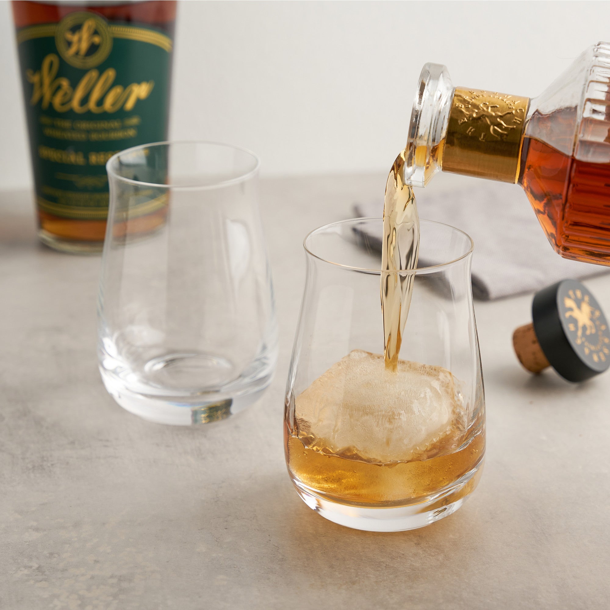 Load image into Gallery viewer, Whiskey being poured into a glass with ice, with a bottle of Weller whiskey in the background.
